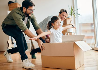 Happy little girl with arms outstretched having fun being pushed by her parents in carton box at new home